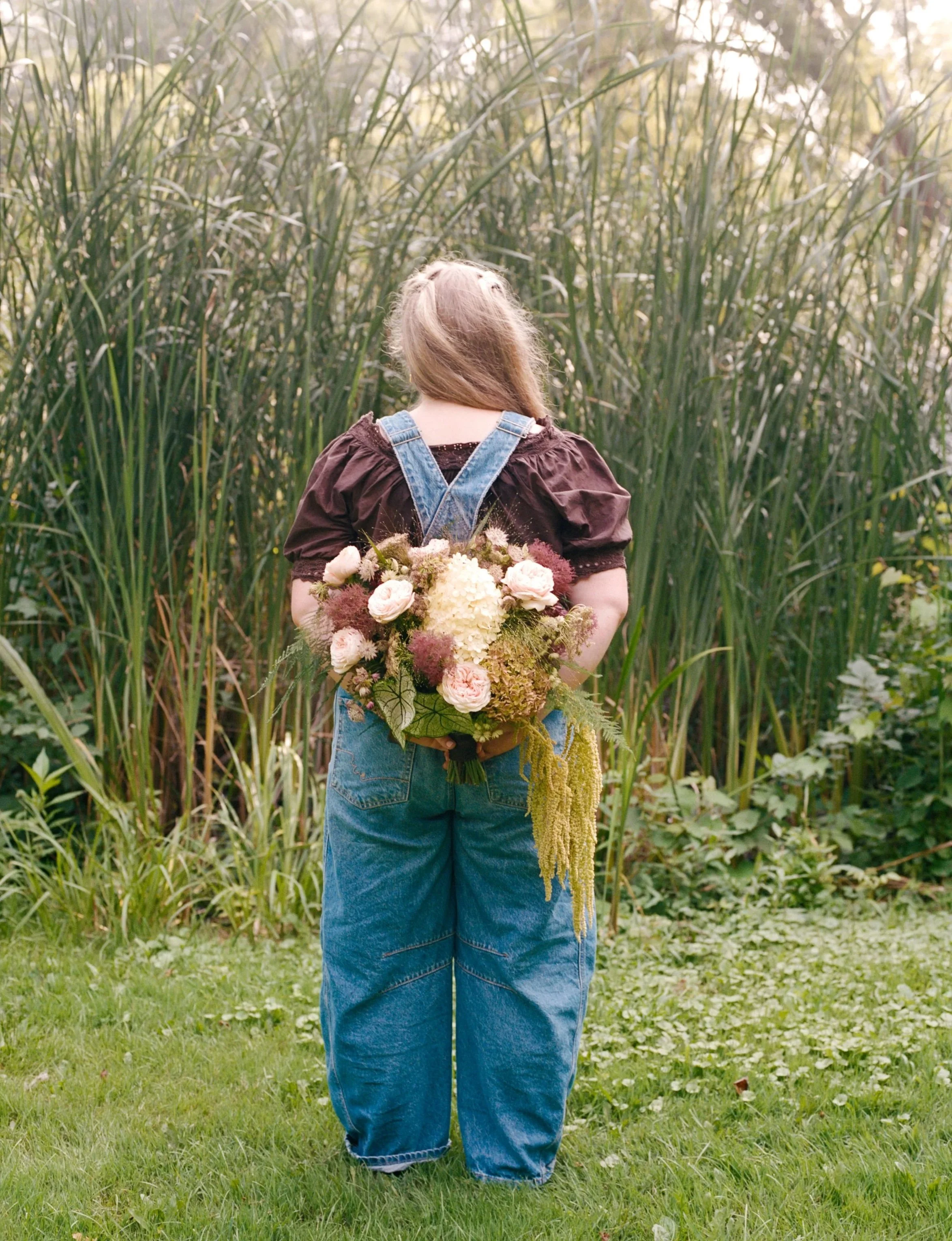 A person standing outdoors in a natural setting, holding a large bouquet of flowers behind their back. The person's face is not visible, and they are wearing a dark shirt and blue jeans. They are standing in front of tall grasses and greenery.