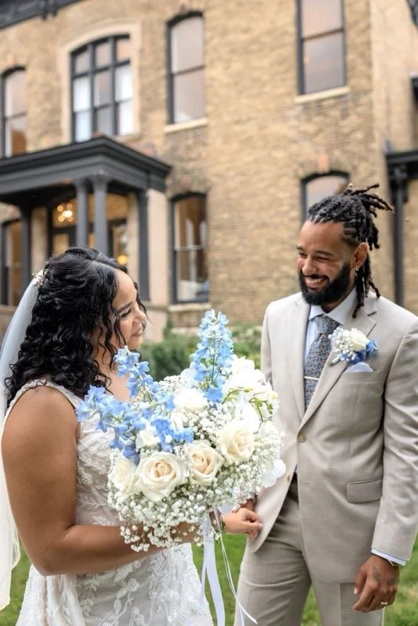 A bride in a wedding dress holding a bouquet of white and blue flowers, smiling at a groom in a light grey suit with a boutonniere, outside in front of a brick building.