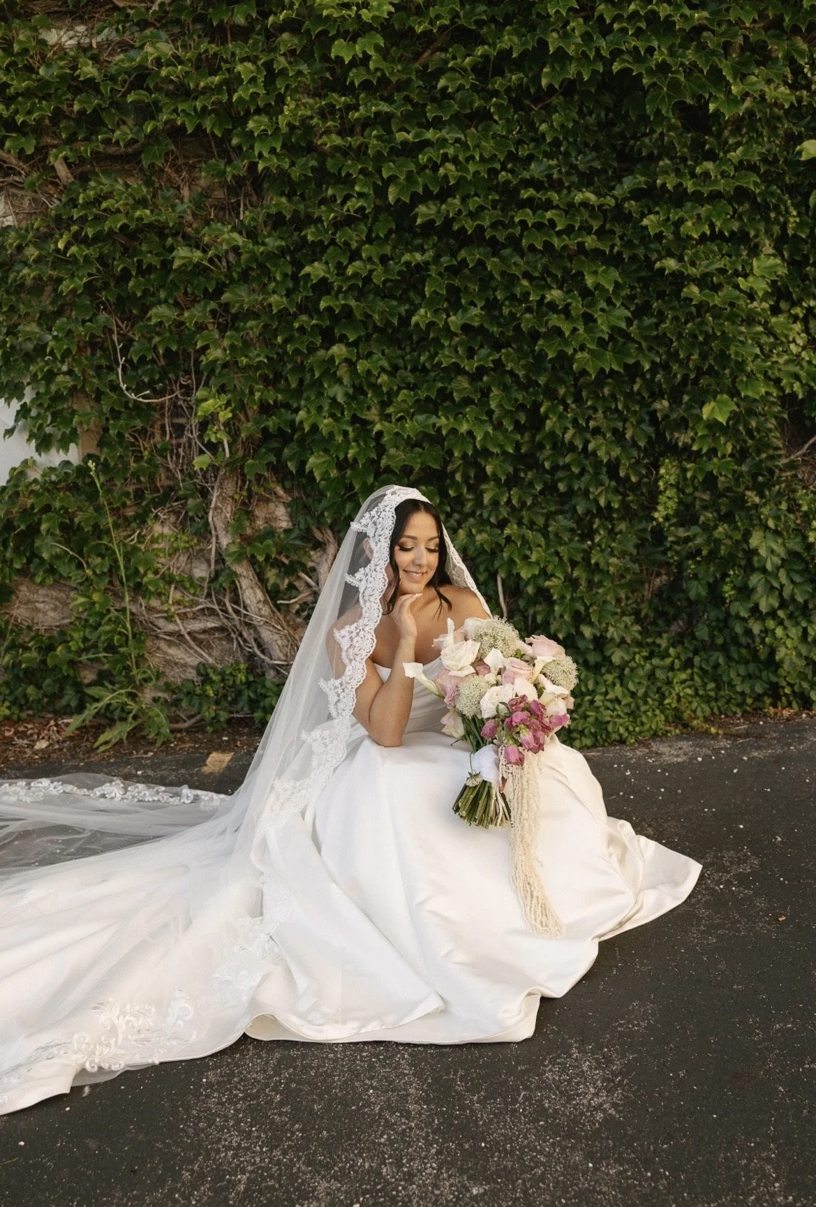A bride in a white wedding gown and lace veil sitting on the ground holding a large bouquet of pink and white flowers, smiling with her eyes closed in front of a lush green ivy-covered wall.