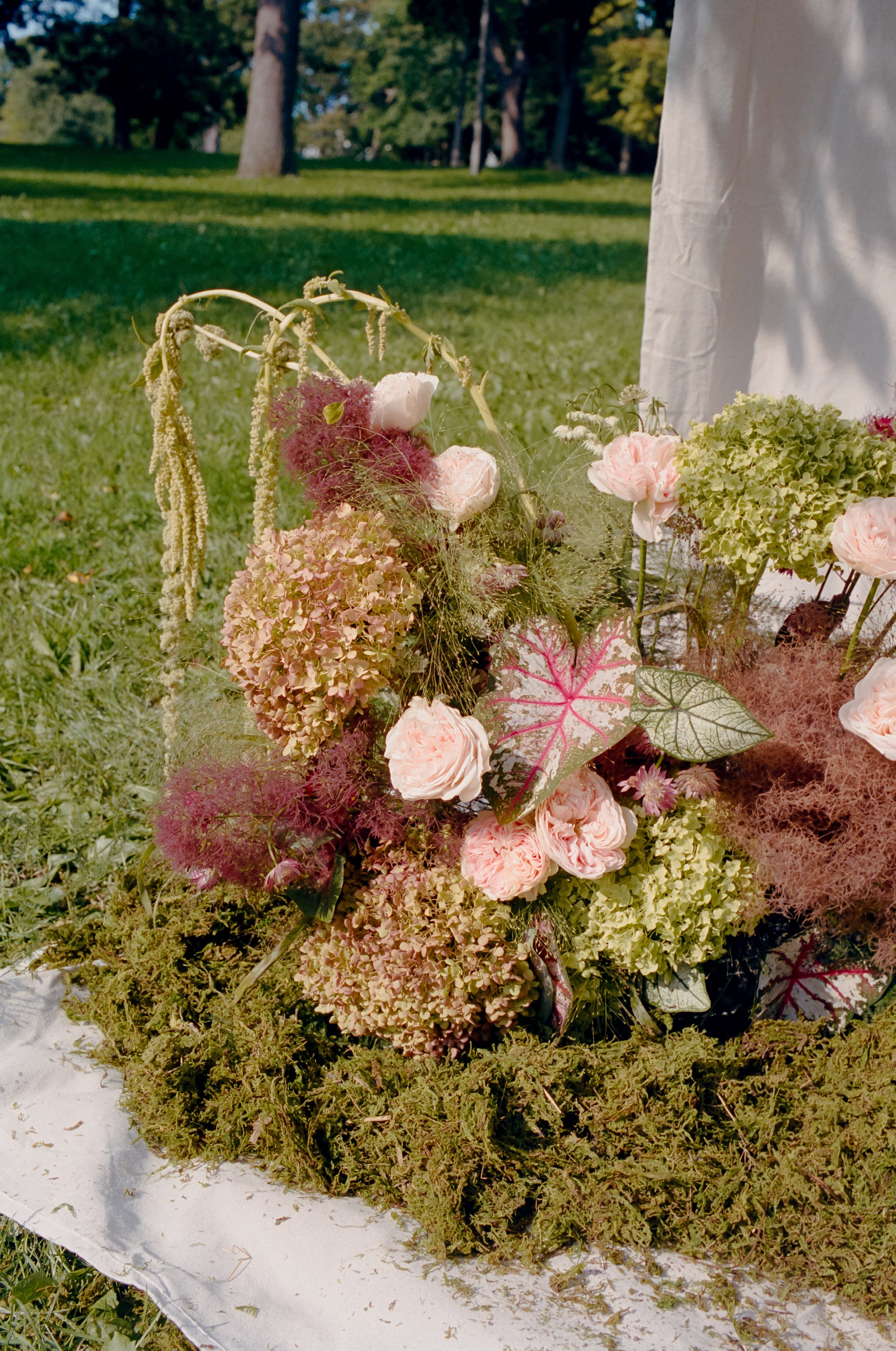 A floral arrangement on a white cloth outdoors, featuring pink roses, green hydrangeas, and various greenery, with a background of grass and trees.