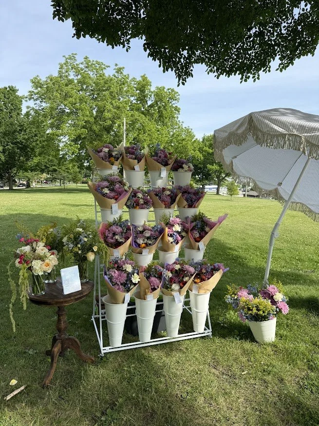 Outdoor flower stand with multiple bouquets of colorful flowers in white cones, a small table with additional flowers, and a white parasol on a grassy field under trees.