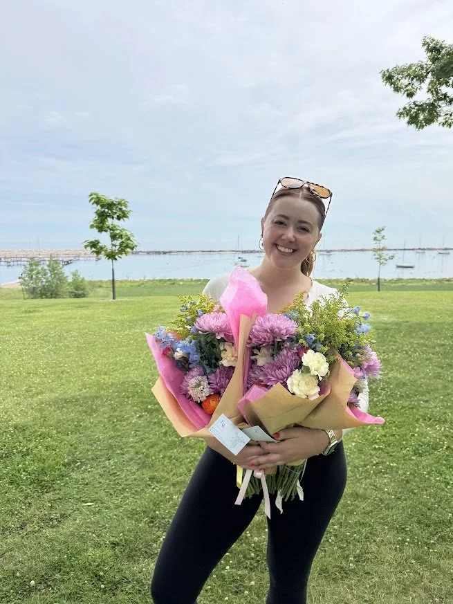 Woman smiling and holding a large bouquet of colorful flowers outdoors near a body of water with trees and a cloudy sky in the background.