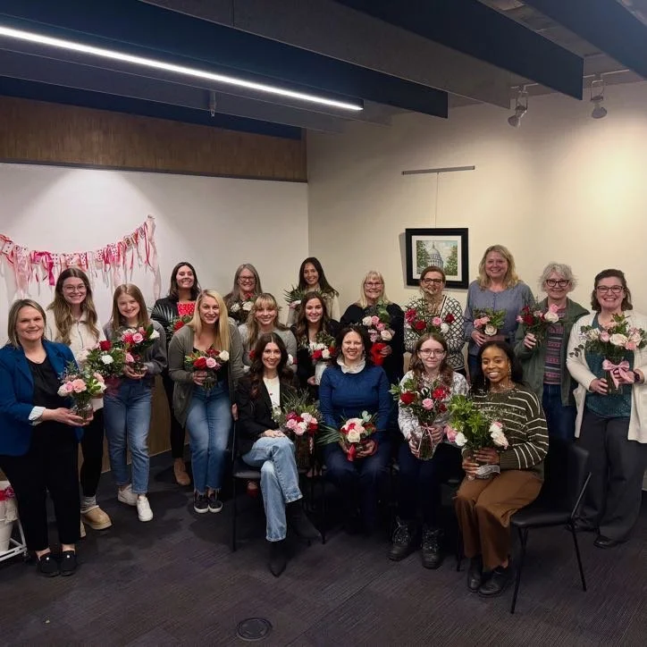 Group of women gathered indoors, holding colorful flower bouquets, smiling for a celebration or event, with pink decorations on the wall and a framed picture in the background.