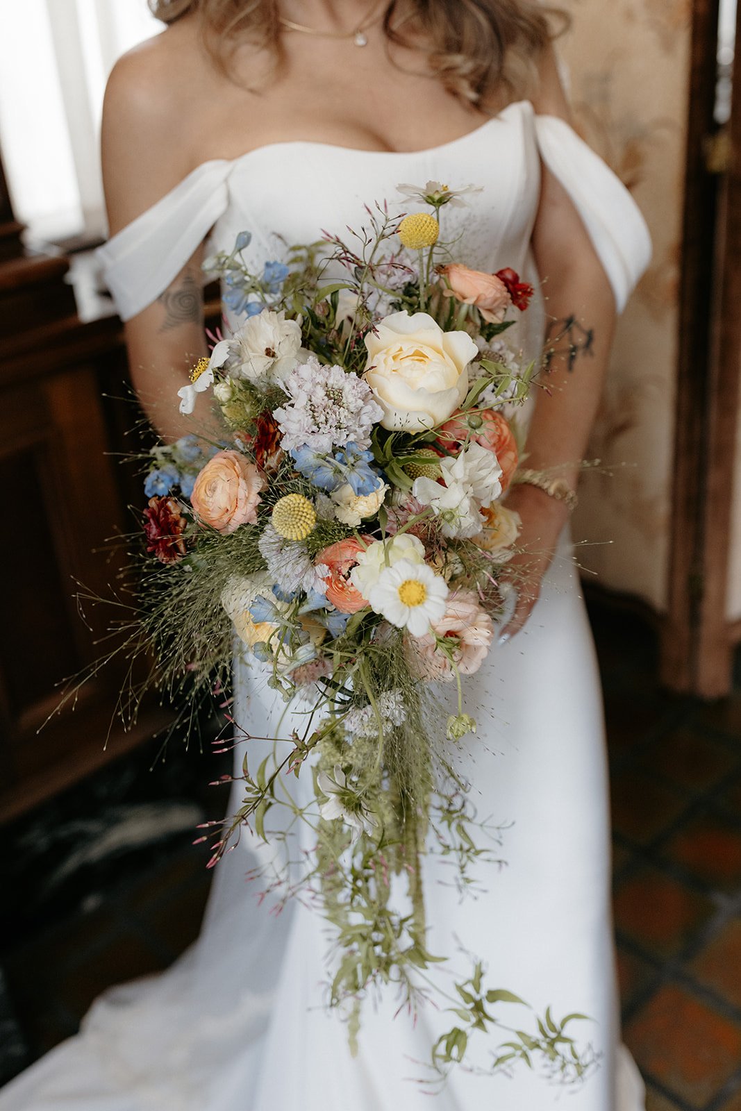 Woman in white wedding dress holding a cascading bouquet of mixed flowers including peonies, daisies, and other blooms.