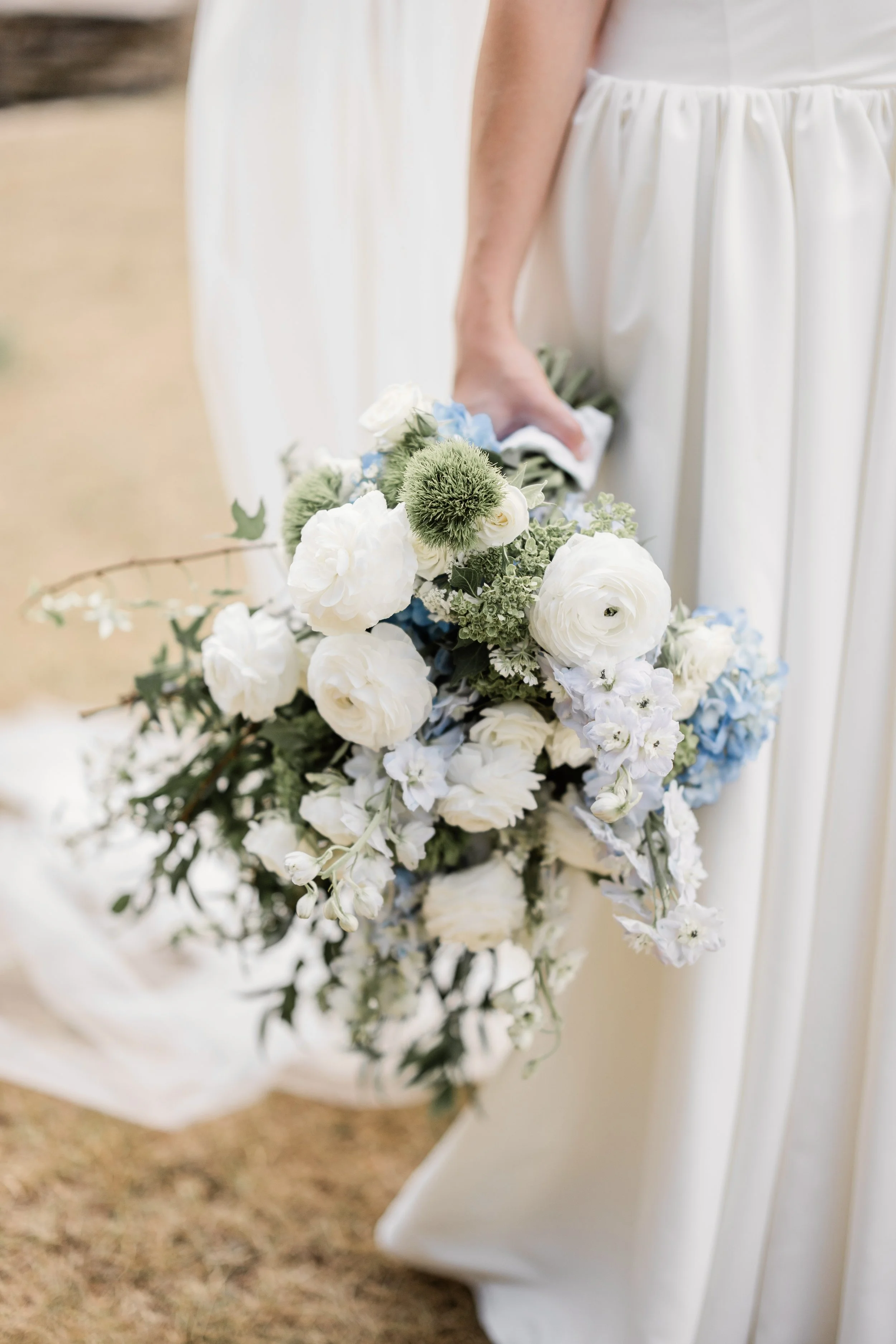 Close-up of a person in a white dress holding a cascading bouquet of white, cream, and light blue flowers.