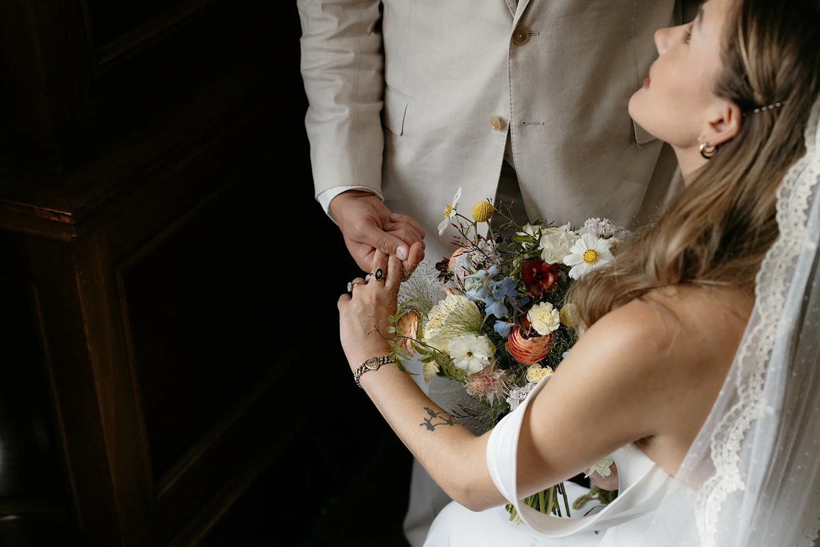 A bride and groom holding hands during a wedding ceremony, with the bride holding a bouquet of flowers and the groom wearing a light-colored suit.