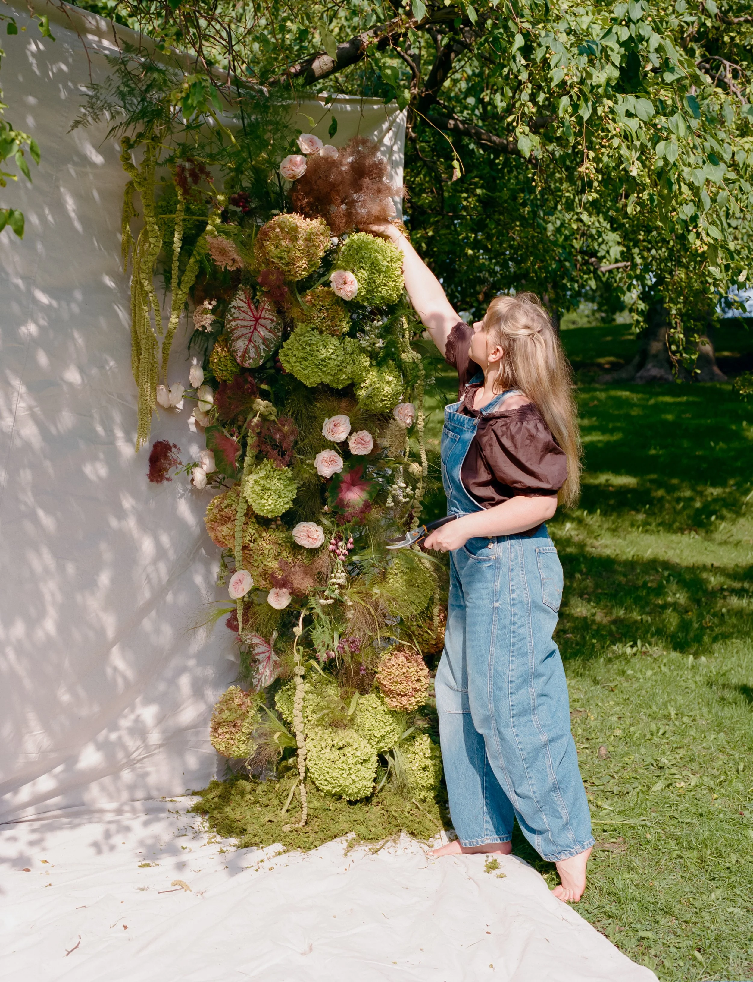 A young girl with long blond hair, wearing denim overalls and a brown jacket, stands outdoors on grass, reaching up to touch a large, colorful floral arrangement on a white wall with greenery in the background.