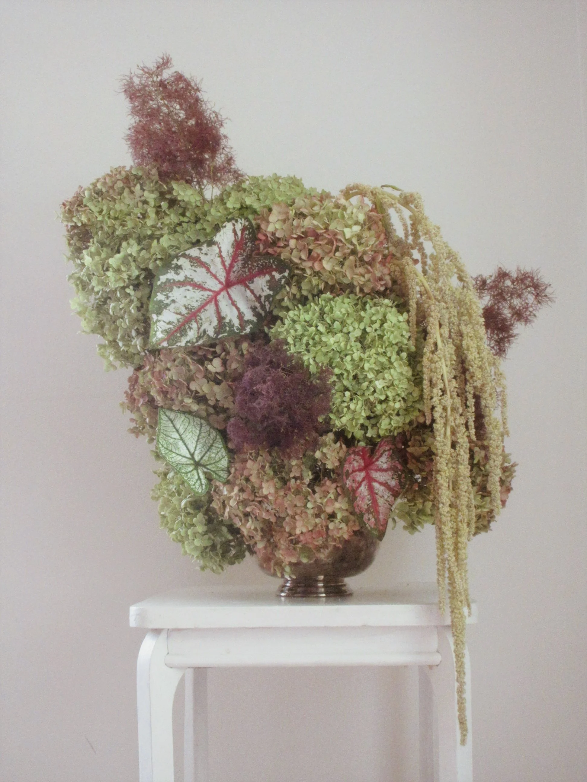 A flower arrangement consisting of various dried green, pink, and purple hydrangeas, with red and green variegated leaves and trailing yellow and purple dried flowers, placed in a small, ornate metal vase on a white table against a plain light-colore