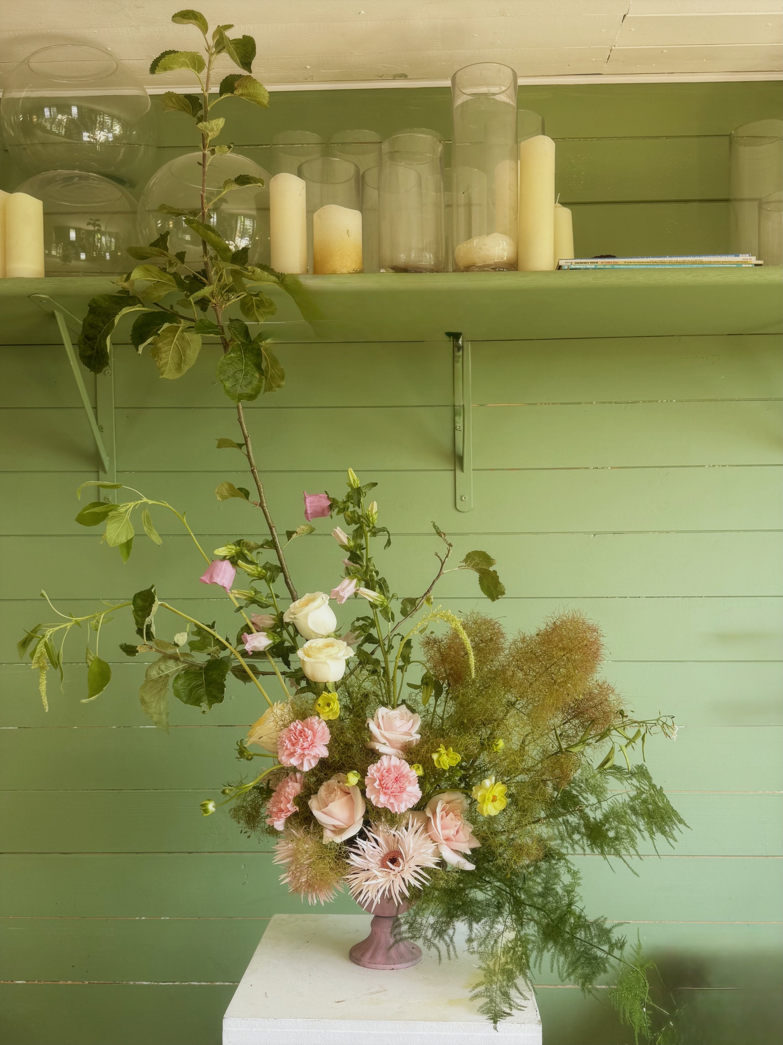 A flower arrangement in a pink vase with various pink, yellow, and white flowers, and green foliage, set on a white stand against a light green wooden wall.