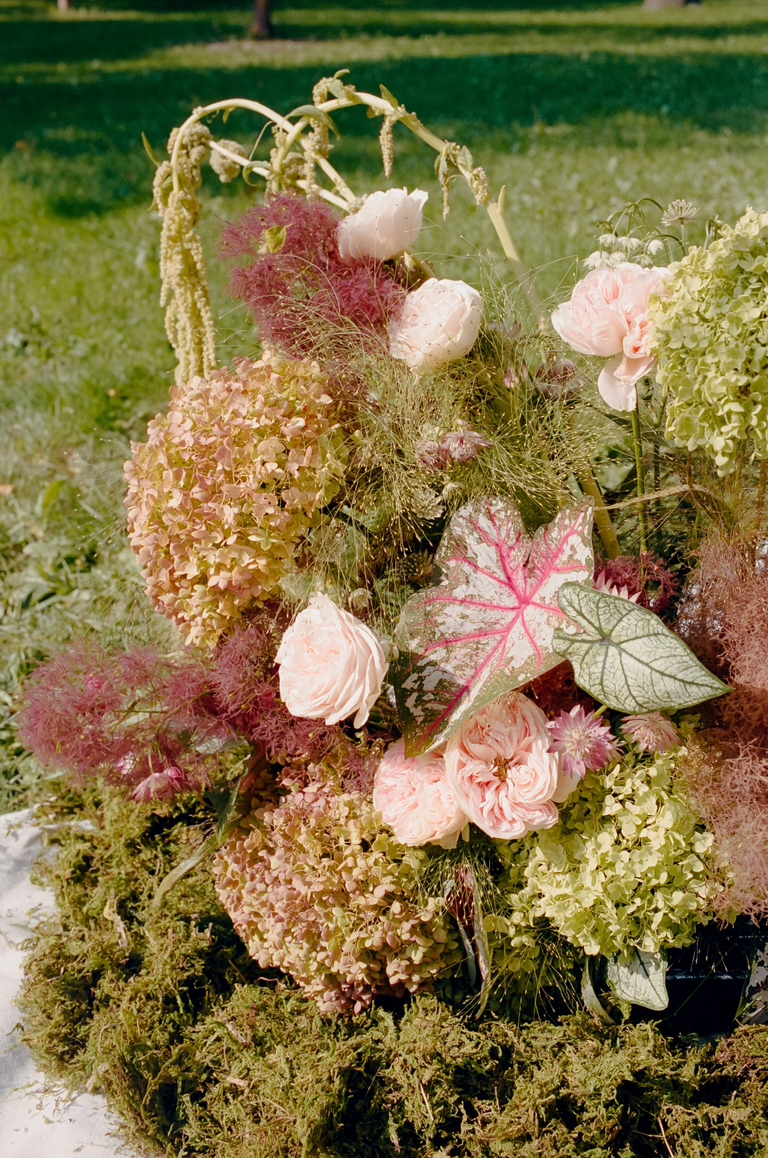 A floral arrangement consisting of pink peonies, hydrangeas, and various leafy plants placed on grass.