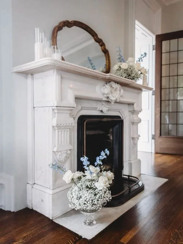 White marble fireplace with a gold-framed mirror on top, decorated with candles and floral arrangements, in a room with hardwood floors and a wooden door with glass panels.