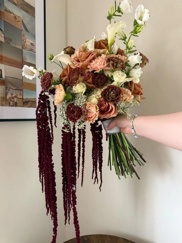 A person holding a bouquet of mixed flowers, including white lilies, roses, and dark red hanging amaranthus, against a cream-colored wall with framed art.
