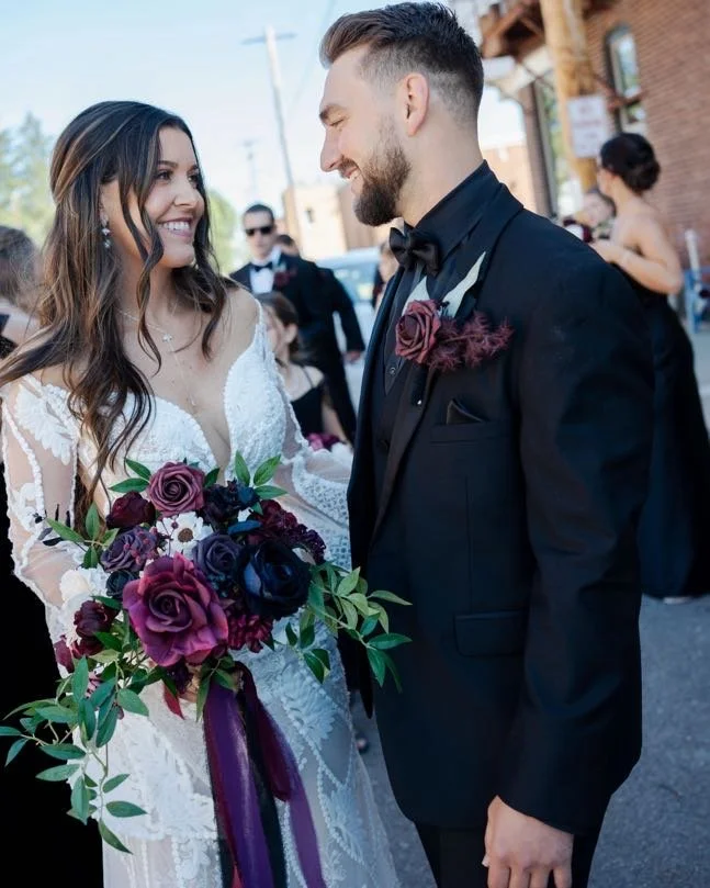 A bride and groom are smiling and looking into each other's eyes outdoors, with the bride holding a colorful bouquet of purple, pink, and dark red roses. The bride wears a white lace dress, and the groom is in a black tuxedo with a black bow tie and 