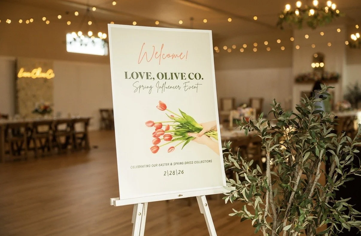 Event welcome sign for LOVE, OLIVE CO. Spring Influencer Event, with a bouquet of tulips being held by a hand illustration, in a decorated event space with tables, chairs, string lights, and greenery.