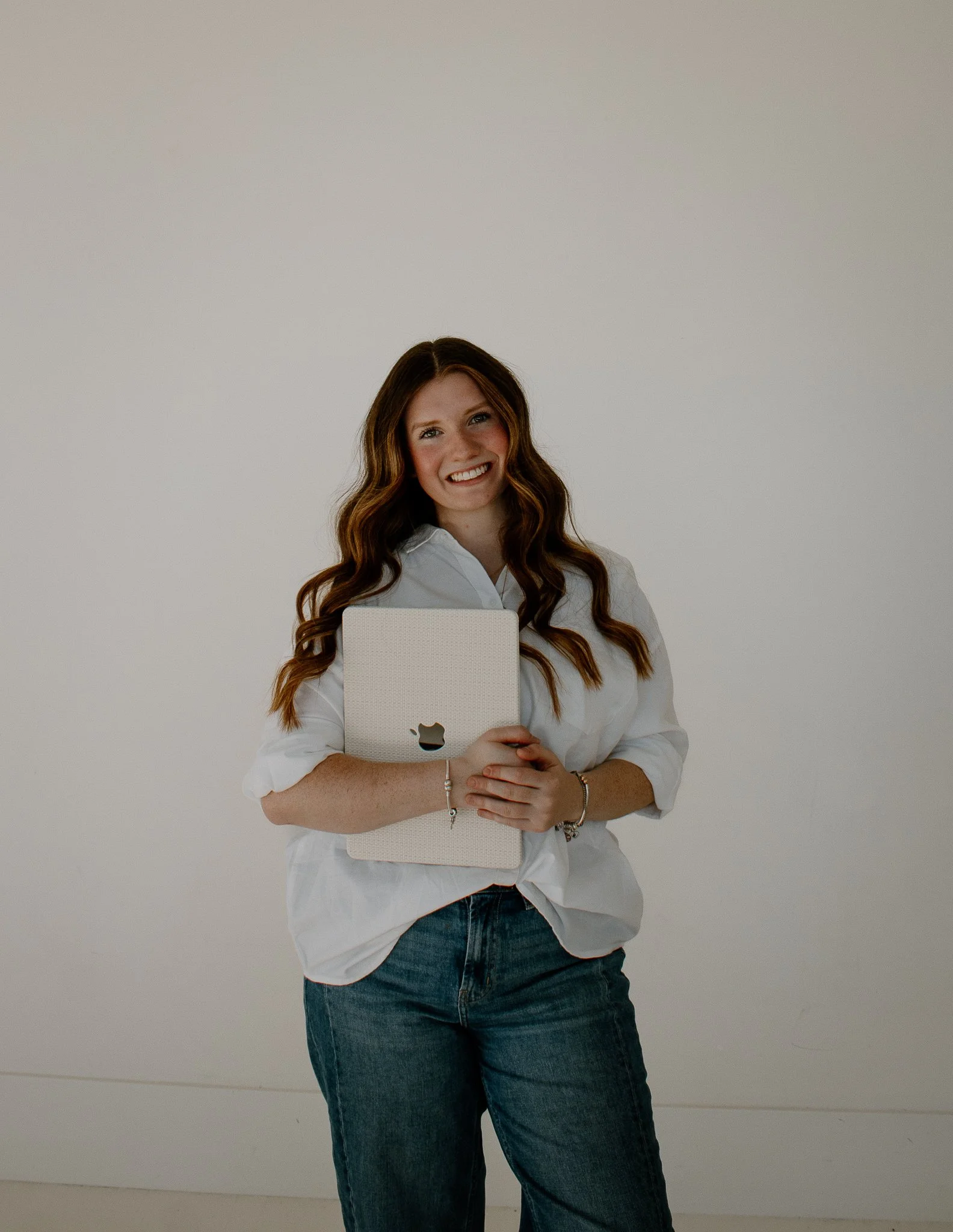A woman with long wavy brown hair wearing a white blouse and jeans holding a silver laptop with a cutout apple logo in front of her, standing in front of a plain white background.