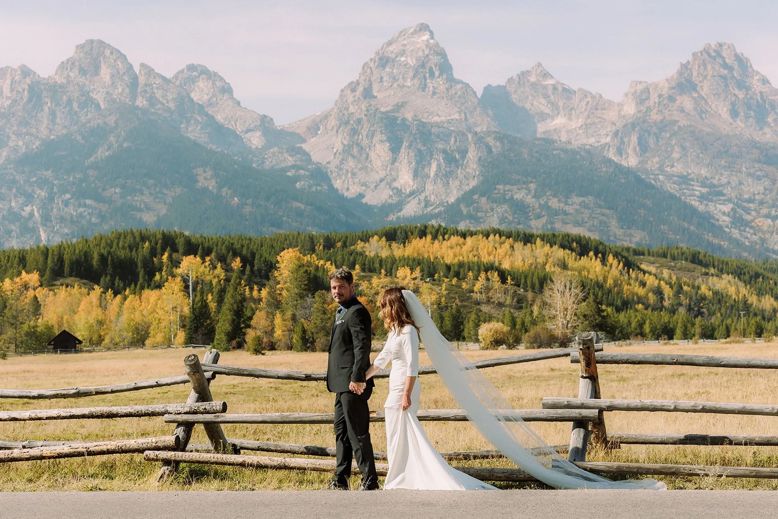 A bride and groom holding hands and standing outdoors in front of a rustic wooden fence, with a mountain landscape in the background displaying tall peaks, green trees, and autumn-colored foliage.