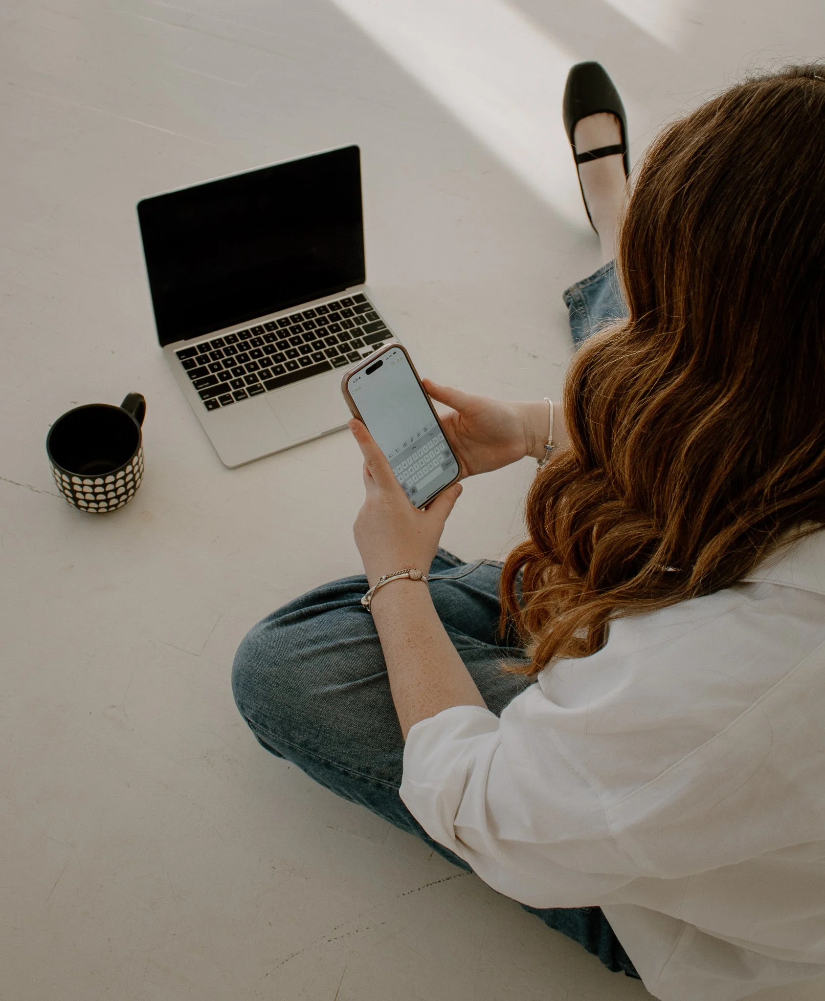 A woman with long brown hair, wearing a white shirt and jeans, sitting on the floor, holding a smartphone, with a laptop and a coffee mug nearby.