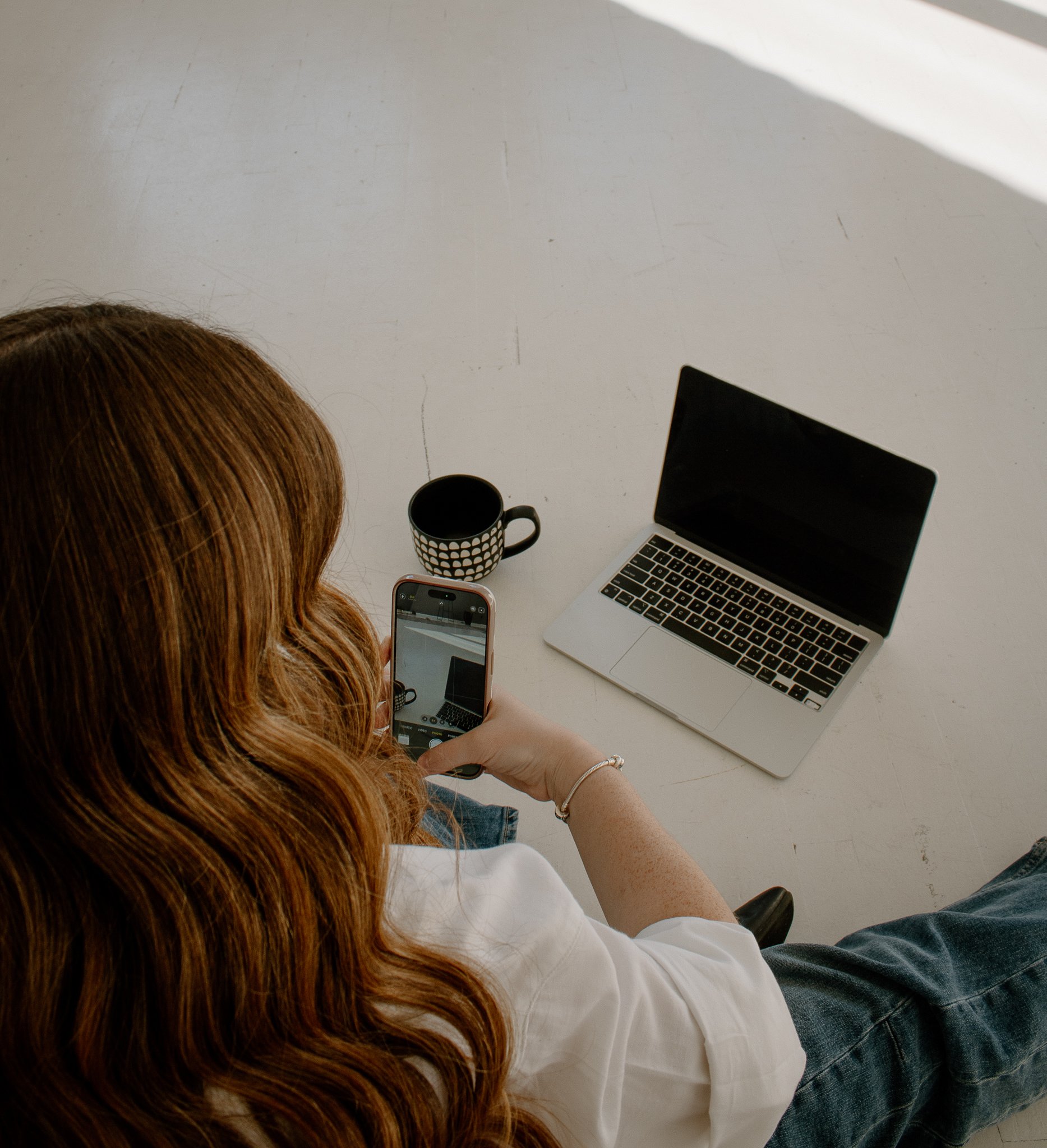 Overhead view of a woman with long red hair sitting at a white table using her phone. On the table are a silver laptop, a black and white polka dot mug, and a black monitor.