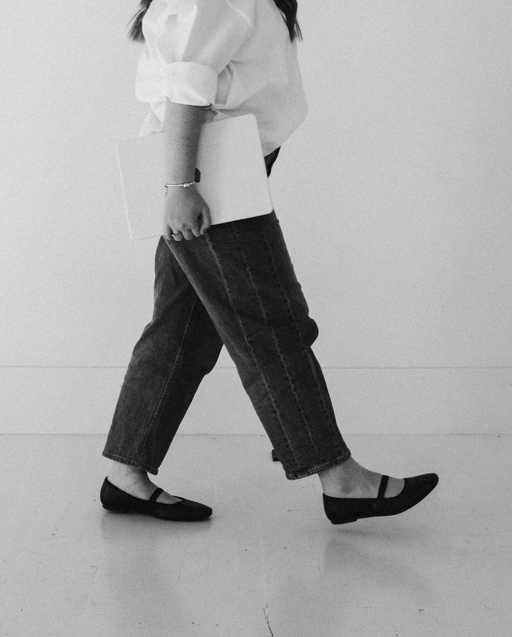 Black and white photo of a woman standing with her legs apart, wearing a white blouse, dark wide-leg jeans, and black Mary Jane shoes. She is holding a rectangular clutch in her left hand and appears to be against a plain wall or floor.