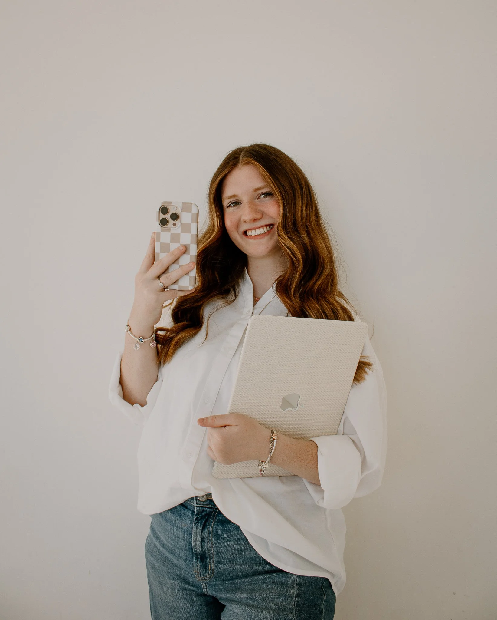 A young woman with long red hair and a white shirt is smiling while holding a smartphone in one hand and an open beige folder in the other. She is standing against a plain off-white wall.