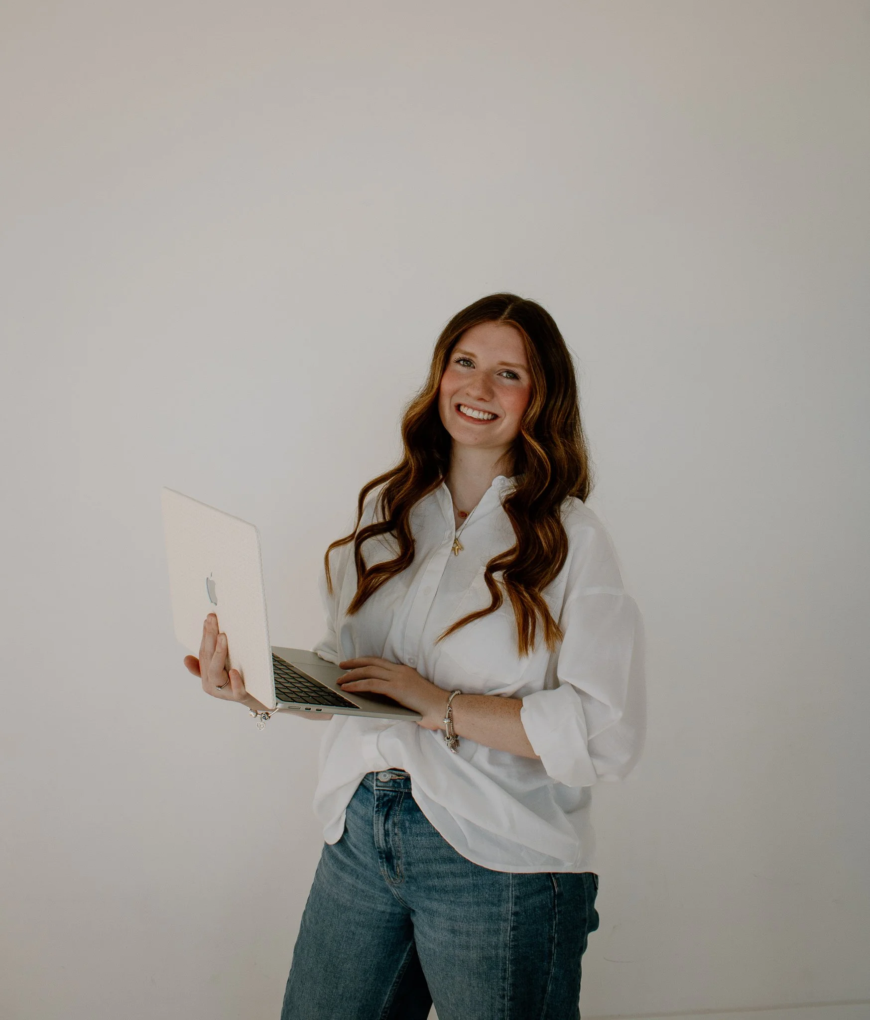 A woman with long, wavy brown hair, wearing a white button-down shirt and blue jeans, smiling while holding an open silver laptop against a plain white wall background.