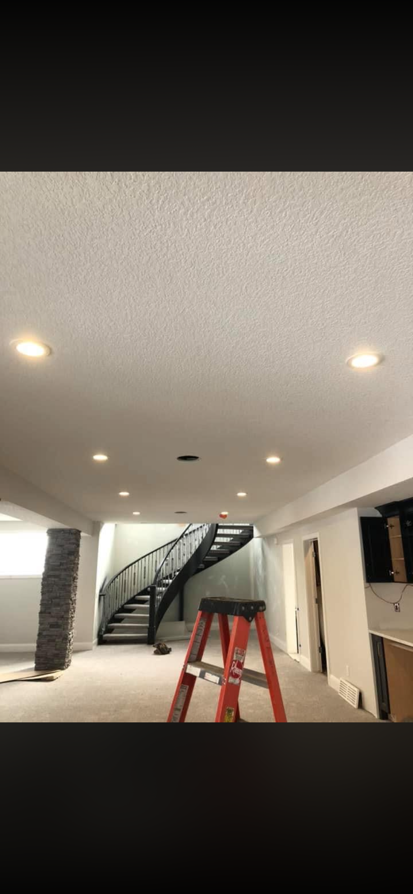 Interior of a house under construction, with a black staircase, a red stepladder, and recessed lighting in the ceiling.