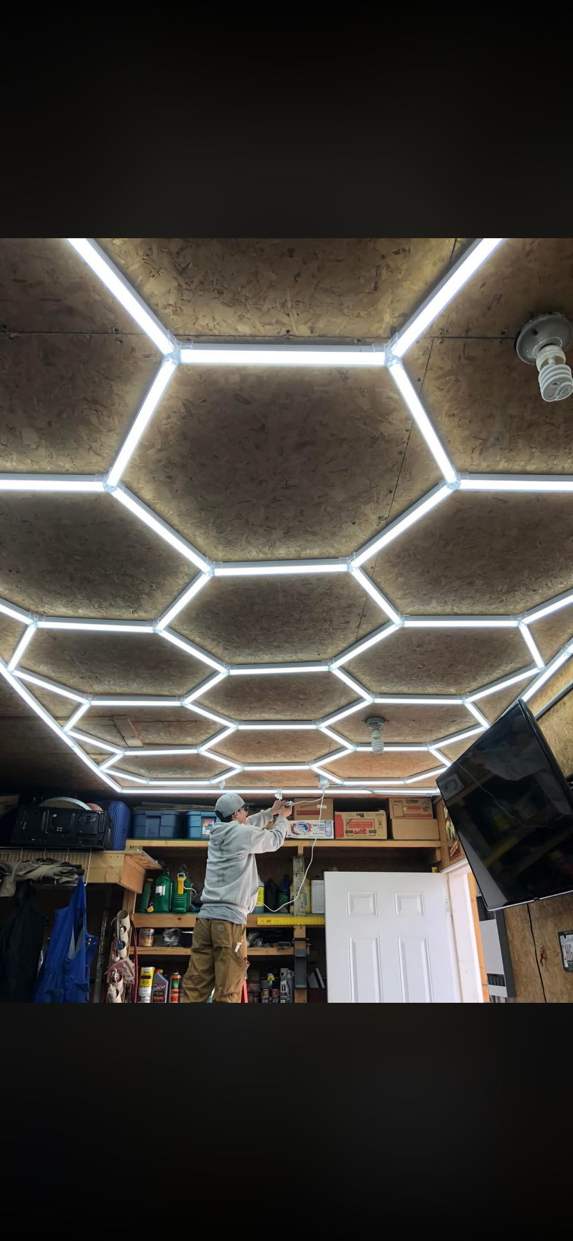 Person installing hexagonal LED ceiling lights in a garage or workshop with wood-paneled ceiling and storage shelves.