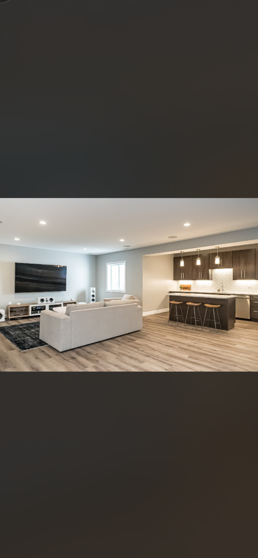 Modern open-concept living room and kitchen with gray walls, hardwood floors, a large flat-screen TV, a light-colored sofa, and a kitchen island with bar stools.