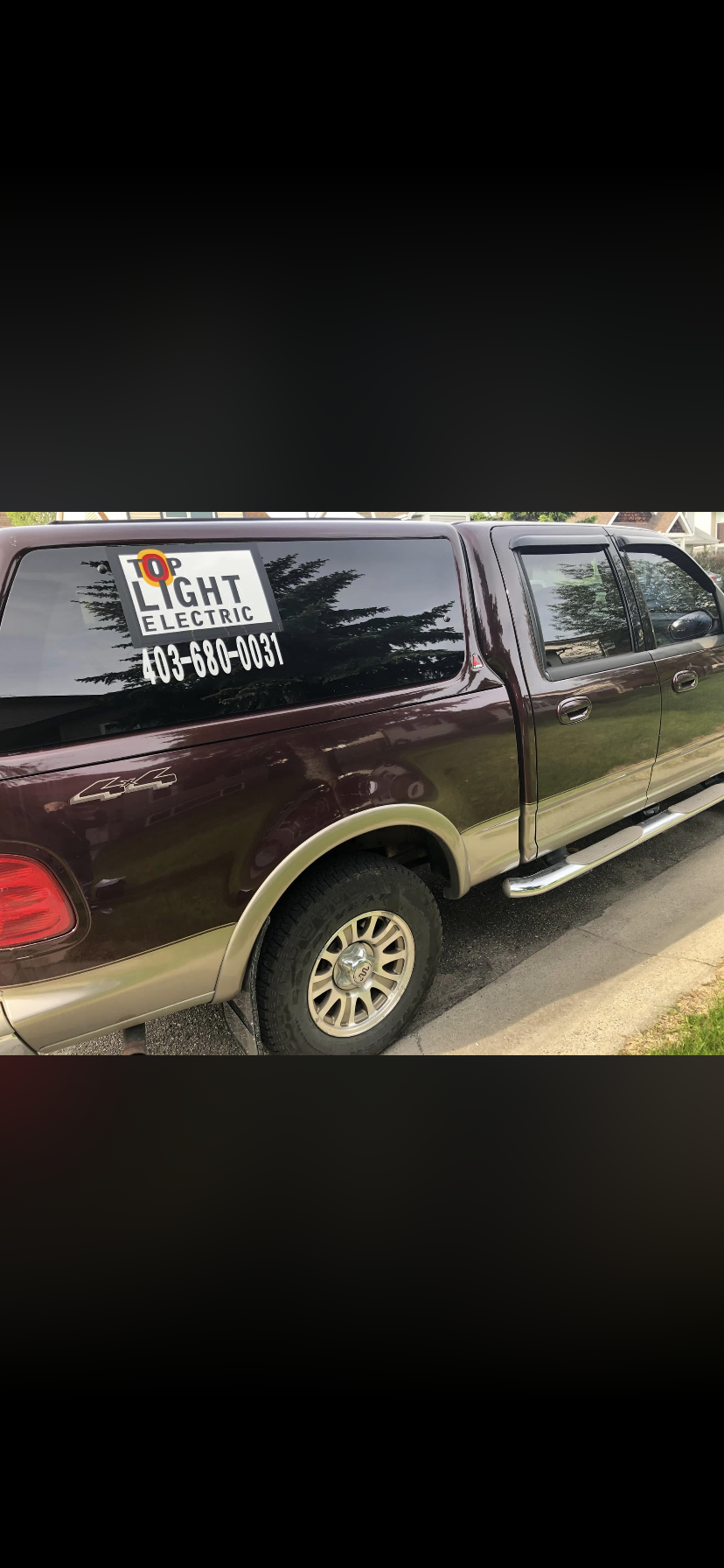 A maroon and beige pickup truck parked on a street with a sign on the side window advertising Top Light Electric and a phone number.