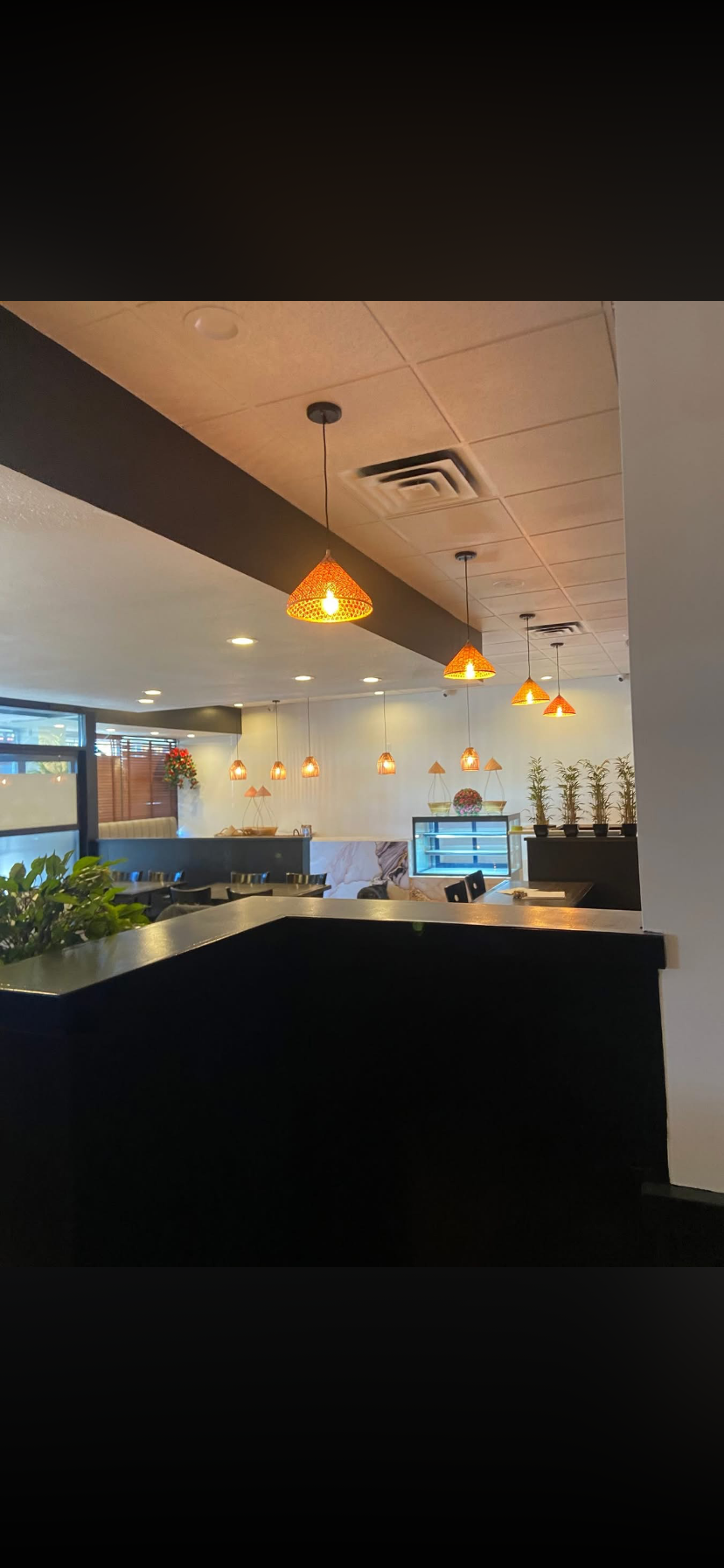 Interior view of a restaurant with hanging orange lamps, black tables, potted plants, and a counter with a glass display case.