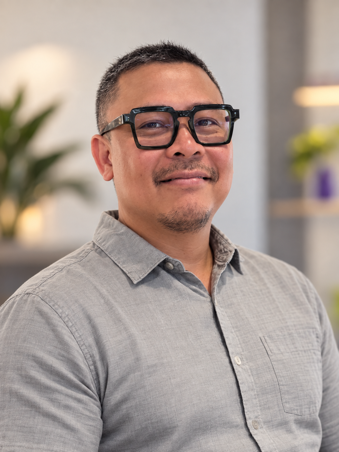 A smiling man wearing glasses and a gray button-up shirt indoors, with blurred plants and decor in the background.