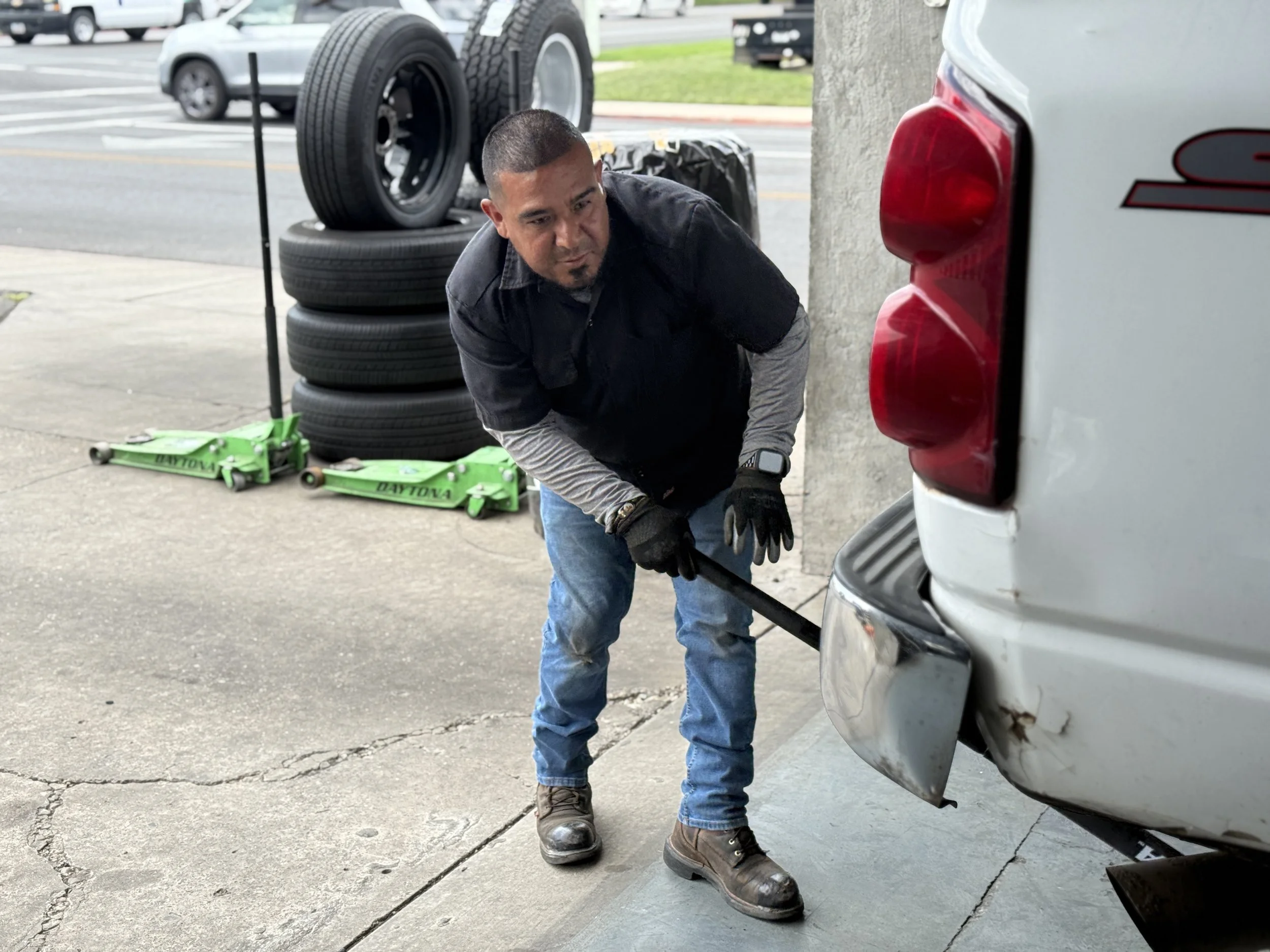 A man is working under the back of a white vehicle, changing a tire and performing maintenance, with a stack of tires, a green hydraulic jack, and various tools around him.