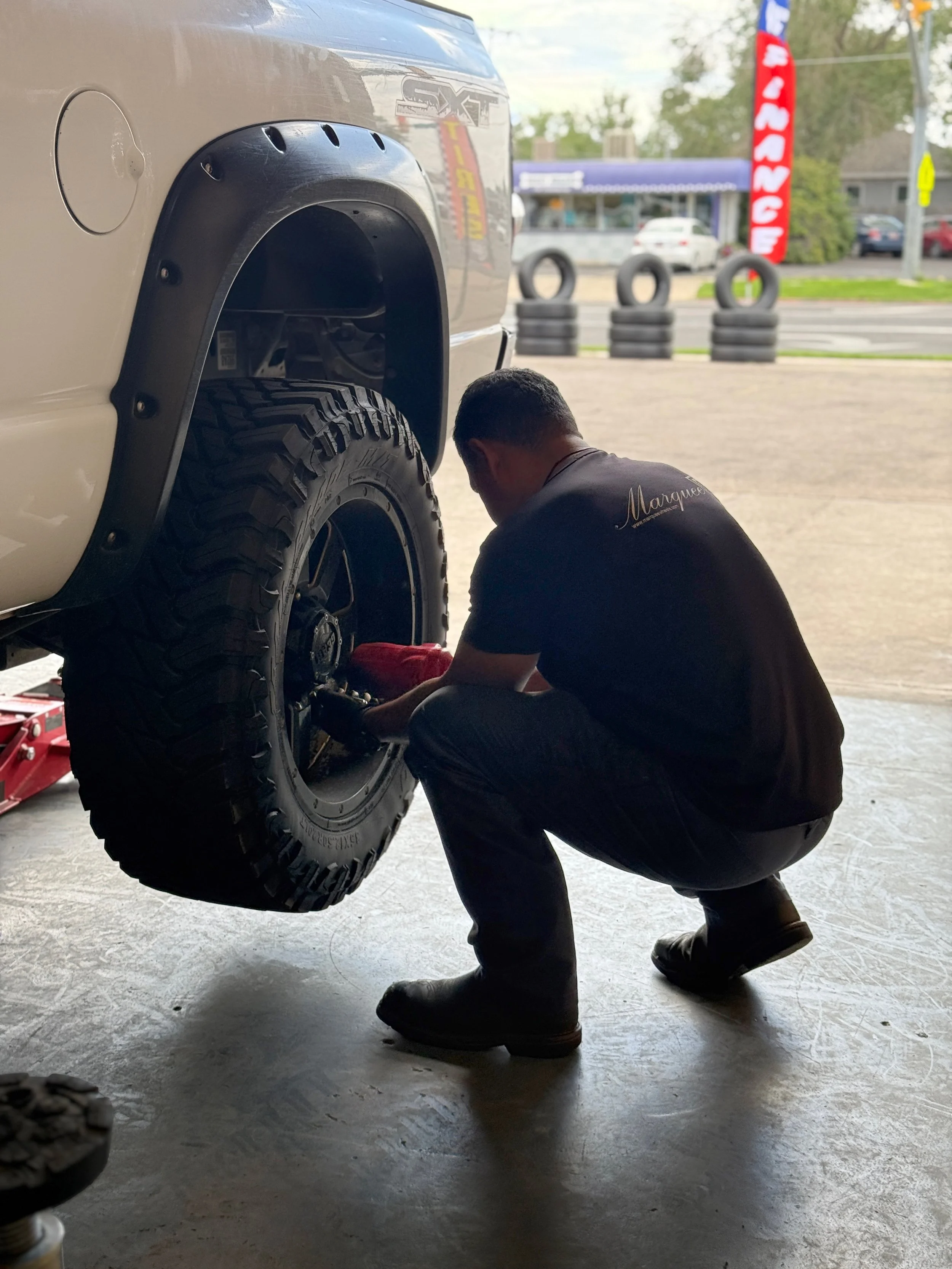 A man is crouched down working on the rear wheel of a beige vehicle in a garage or workshop, with tires and a sign in the background.