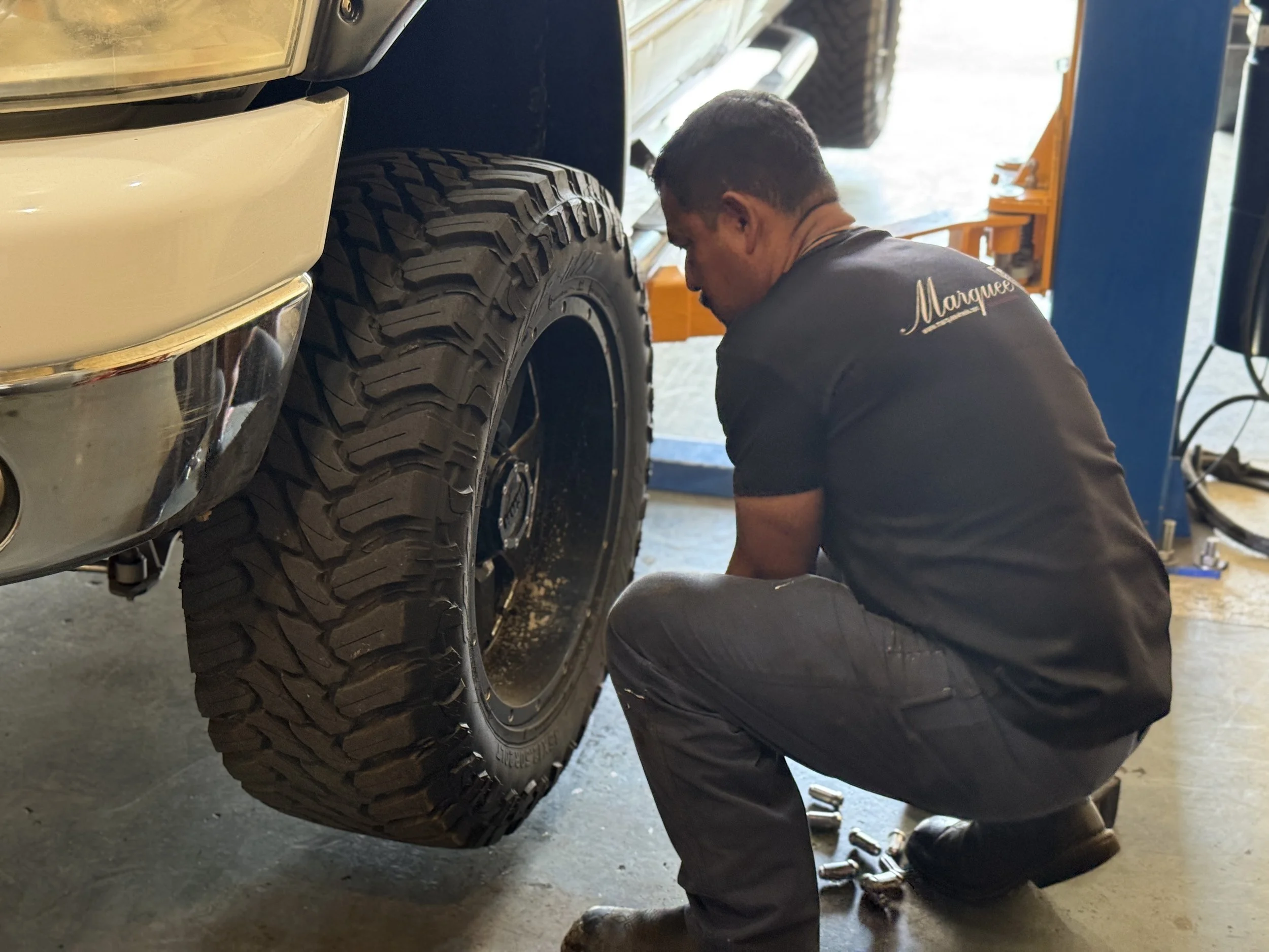 A man working underneath a vehicle with large off-road tires, in a garage or workshop setting.