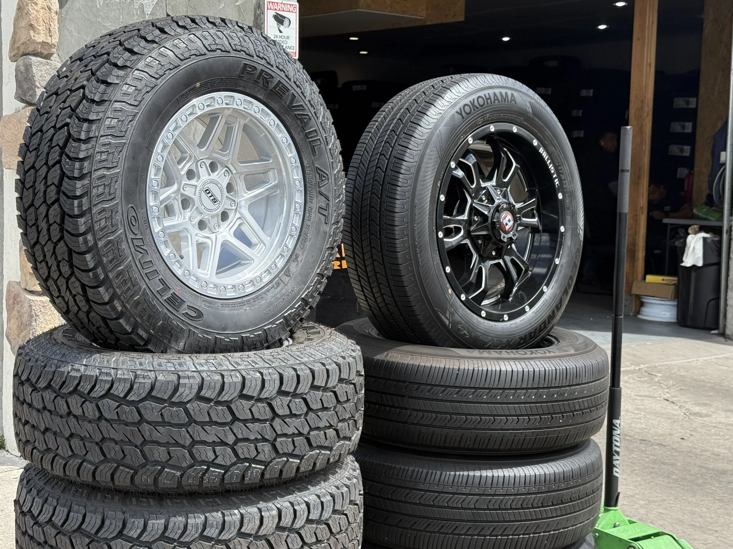 Stacked tires with various tread patterns and brands, displayed at an outdoor tire shop or garage.