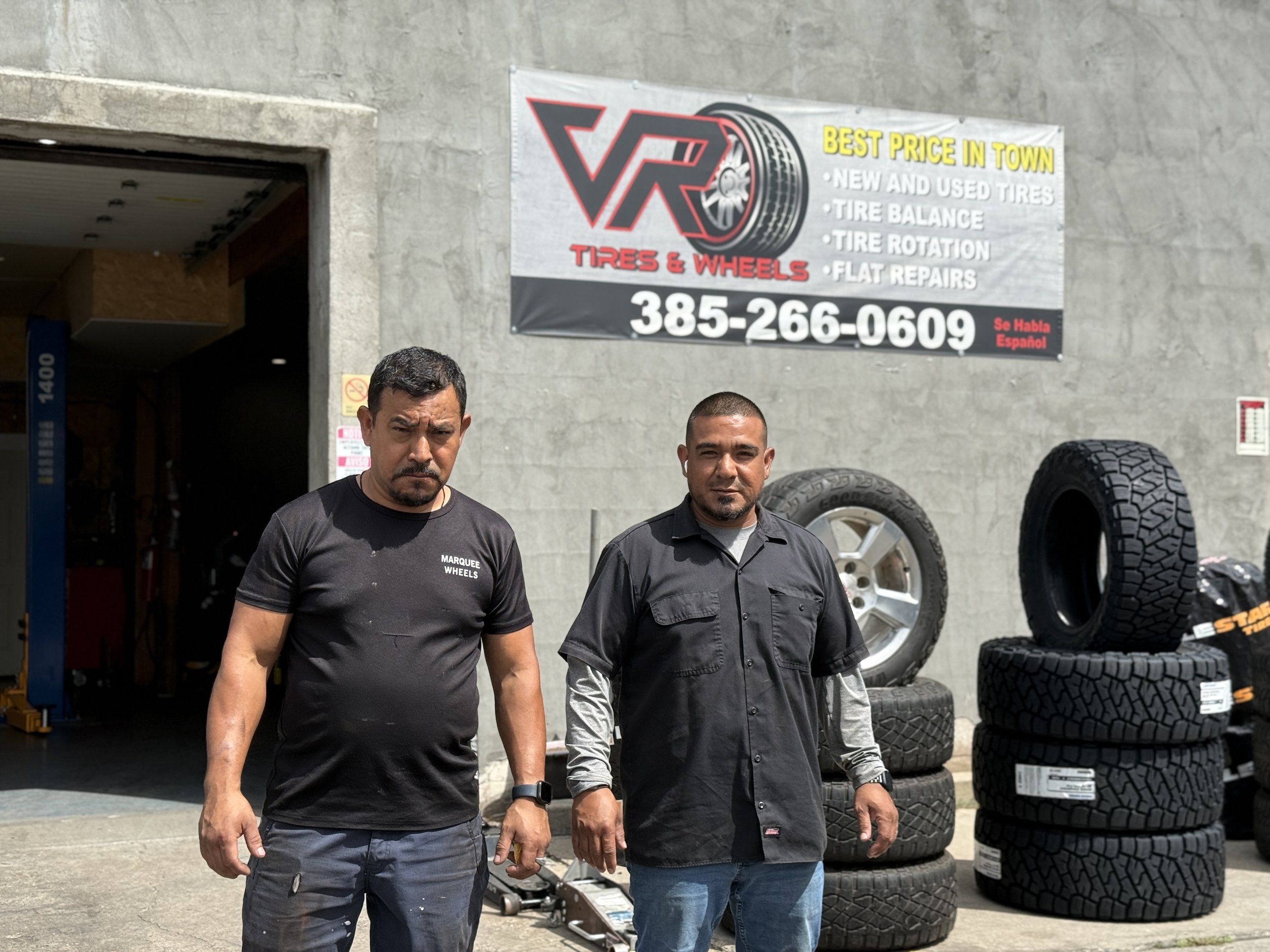 Two men standing outside an automotive tire and wheel shop with a large sign and a stack of tires in the background.
