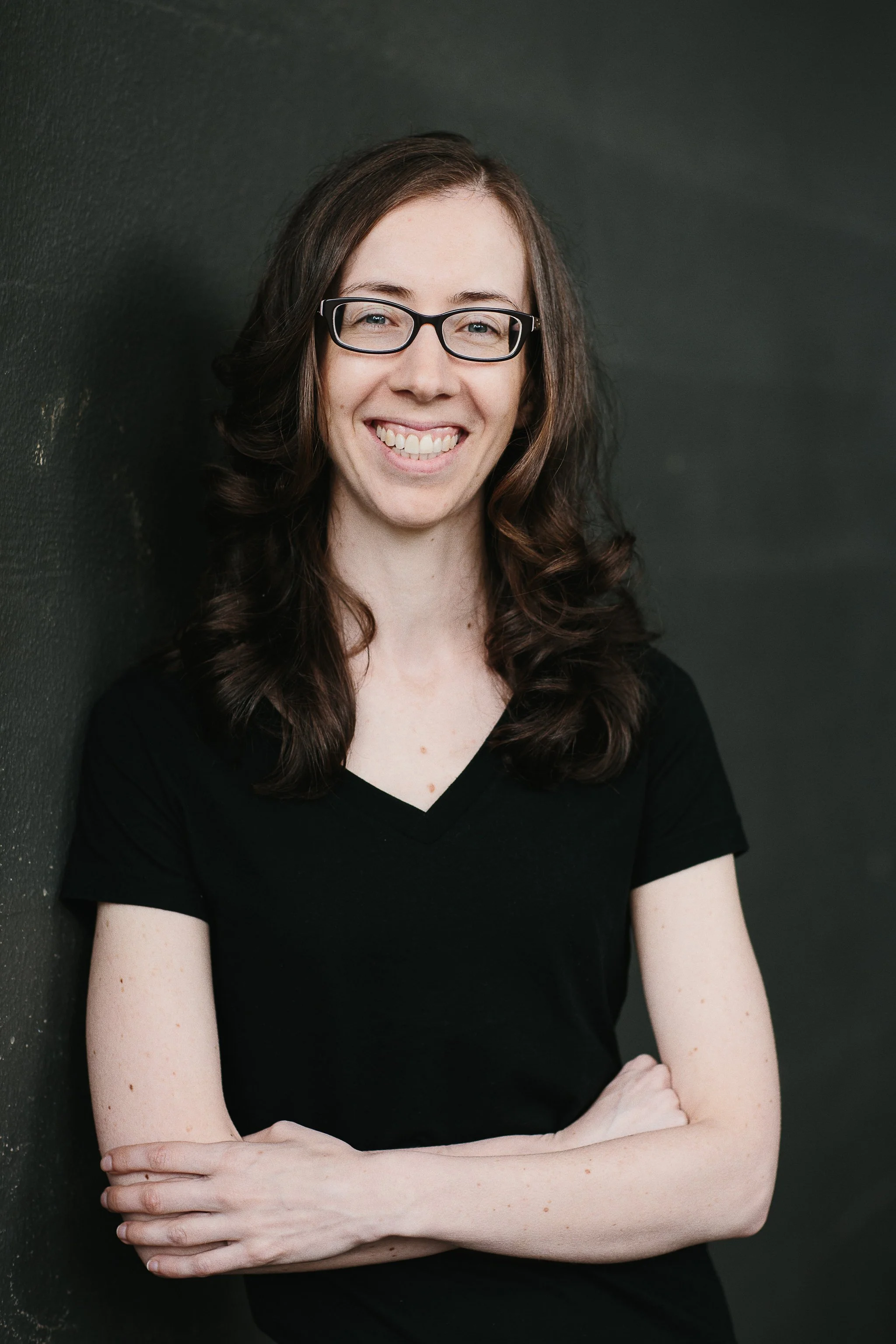 Portrait of a woman with long, dark, curly hair and glasses, smiling, wearing a black V-neck shirt, arms crossed, standing against a dark wall.