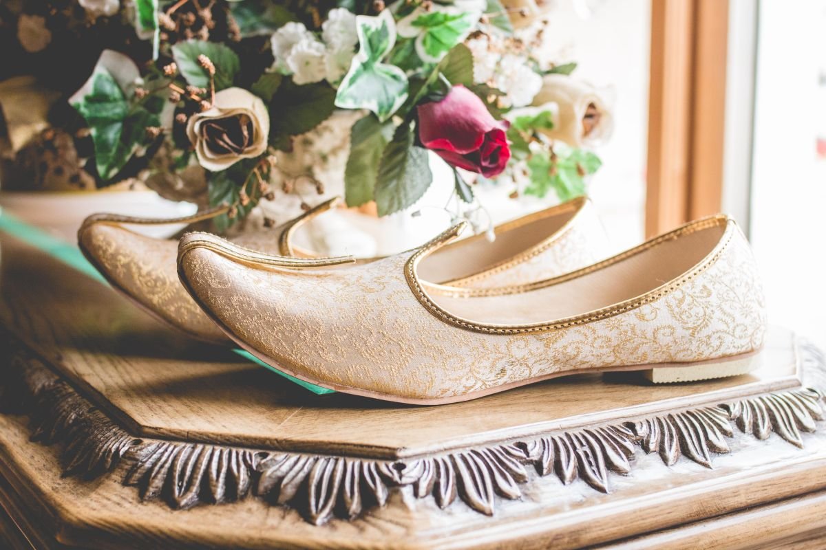 A macro detail shot of a groom's traditional gold embroidered mojari wedding shoes sitting on a decorative wooden table next to a floral arrangement in Winnipeg, Manitoba.