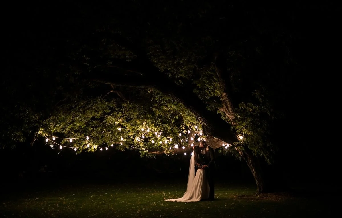 A bride in a white dress and a groom in a dark grey suit stand under an ash tree at night with warm-coloured string lights softly illuminating them at Ashgrove Acres in Manitoba.