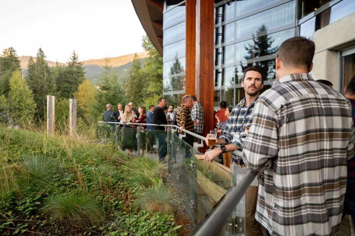 A candid corporate event photograph of business professionals networking on an outdoor glass-railed patio at a business development meeting in Whistler, with mountains and pine trees in the background.