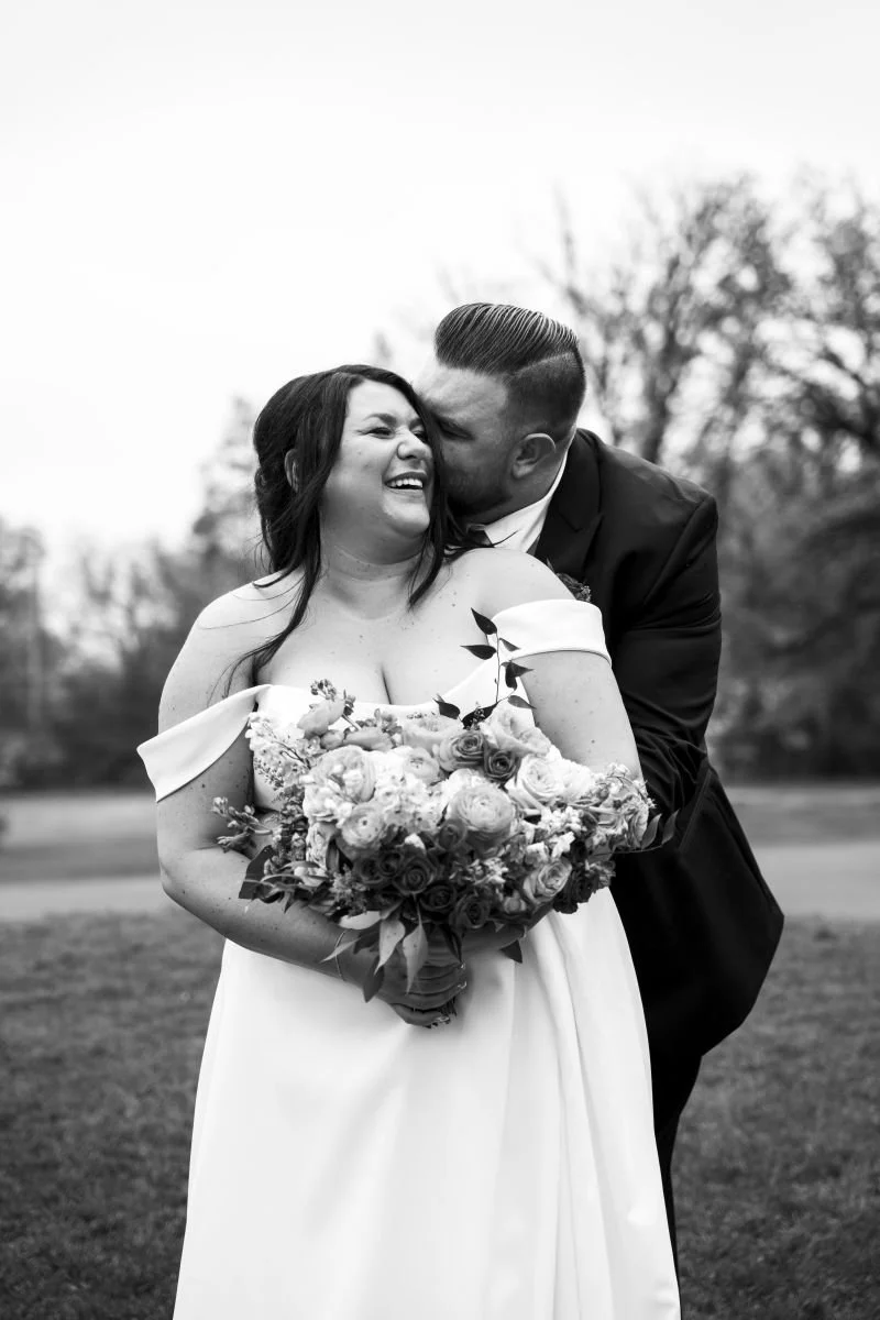 Black and white portrait of a groom embracing his laughing bride from behind during their wedding at St. Boniface Golf Club in Winnipeg.