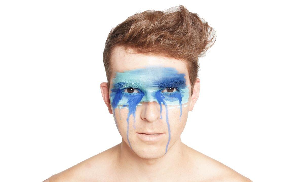 A high-fashion studio beauty portrait of a shirtless young man looking forward against a clean white background, with abstract streaks of blue paint dripping down his face at a photography studio in Winnipeg, Manitoba.