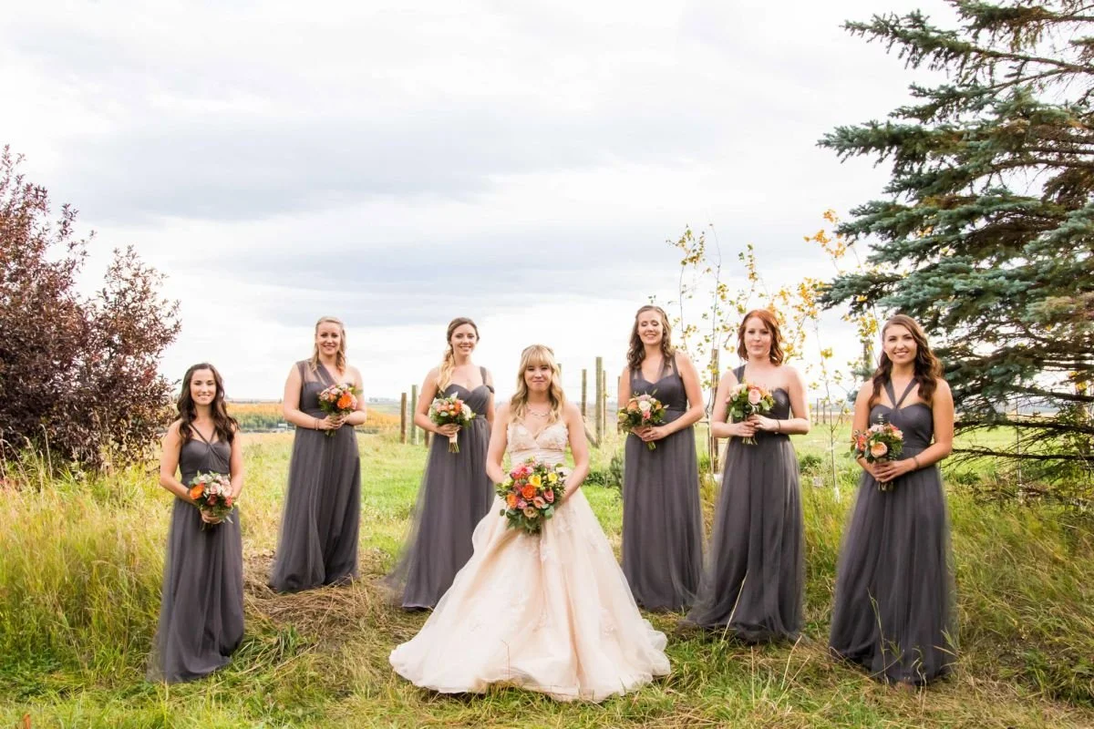 A bride in a beige tulle maxi dress stands with her bridesmaids in charcoal grey maxi dresses holding orange and pink floral bouquets at The Saskatoon Farm venue during an outdoor destination wedding in Foothills County, Alberta, captured by a Winnip