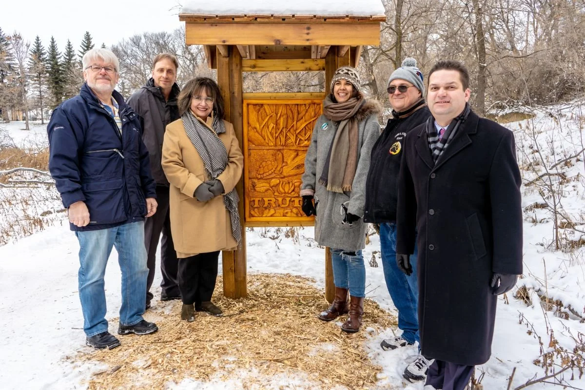 Members of the Winnipeg Arts Council team pose together in winter clothing next to a newly unveiled carved wooden art installation bearing the word Truth in a snow-covered outdoor park setting.
