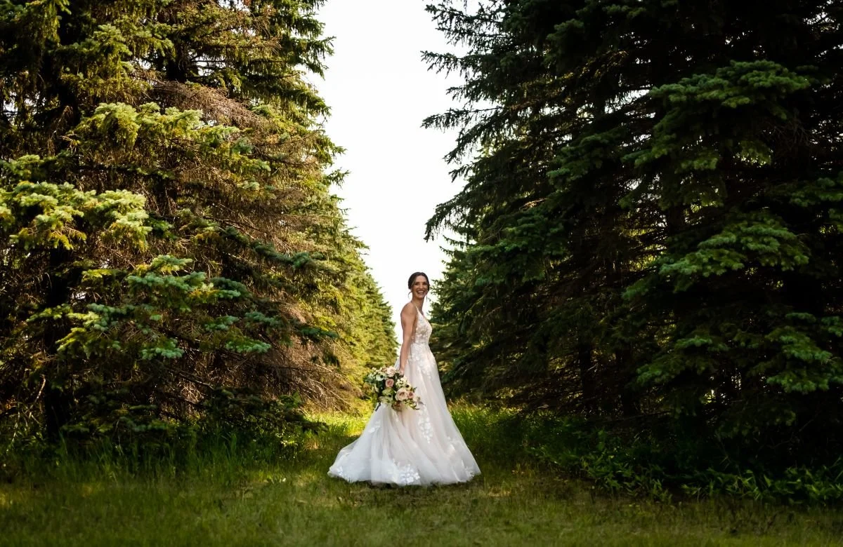 Photo of woman standing sideways, looking at the camera and smiling while she holds her bouquet in a corridor of pine trees.