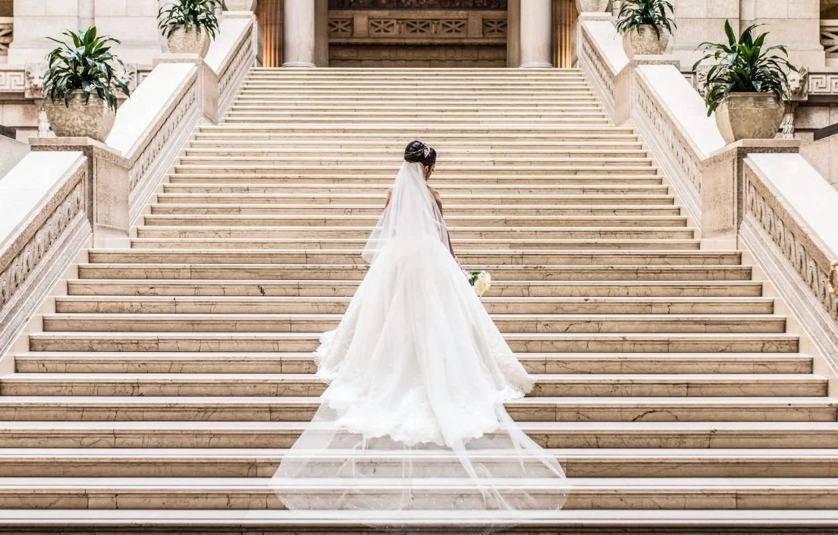 A portfolio preview tile showing a back-angle portrait of a bride in a ballgown with a long sheer veil walking up the grand limestone staircase inside the Manitoba Legislative Building, linking to the full Winnipeg bridal portrait gallery.