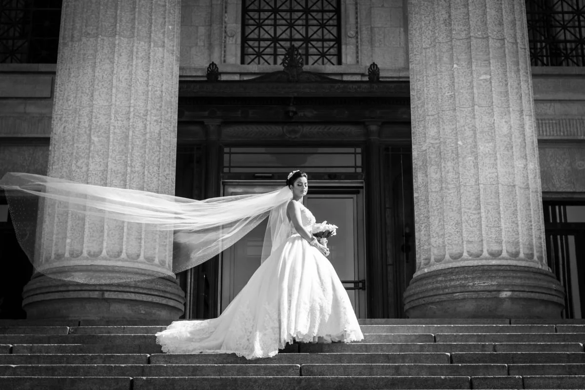 A dramatic black and white wedding portrait of a bride with a long flying veil standing on the steps of the Manitoba Legislative Building, captured by a Winnipeg wedding photographer.