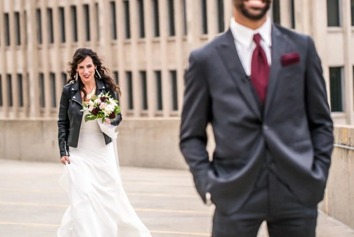 A bride in a white dress and short black leather motorcycle jacket approaches the groom in a dark grey suit with a burgundy tie and pocket square at the top of a parkade in the Exchange District of Winnipeg, Manitoba.