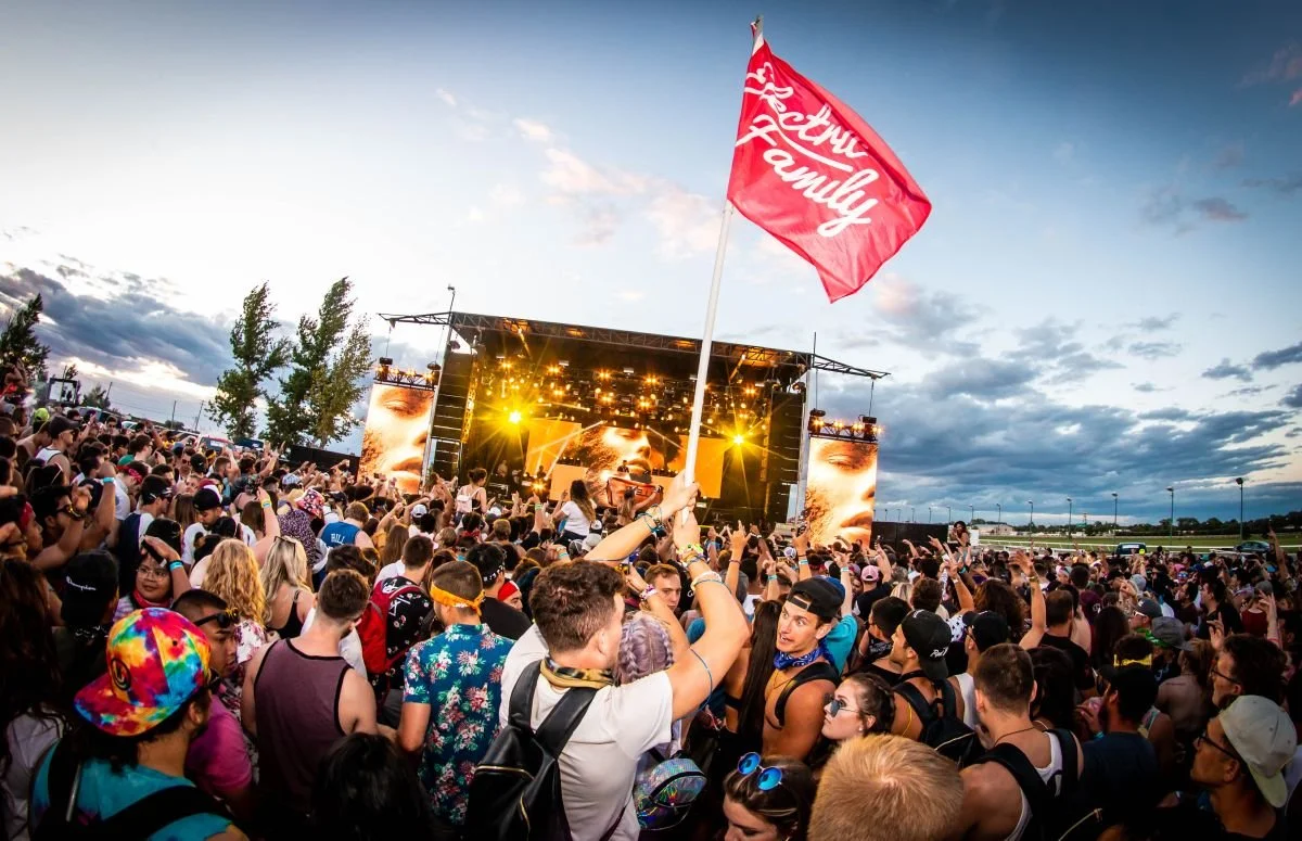 A massive festival crowd watching EDM DJ Illenium perform live at Summer of Sound in Winnipeg, Manitoba while an attendee waves a large Electric Family flag.