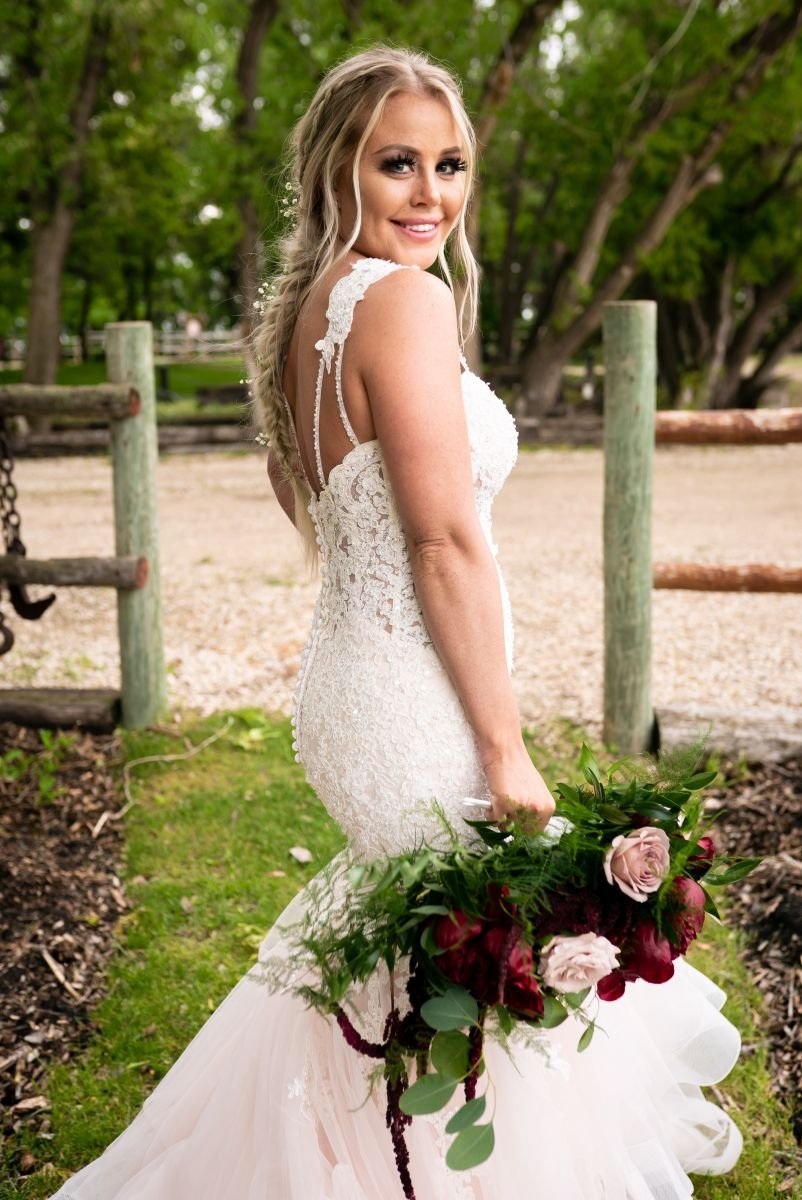 A full-length bridal portrait of a smiling bride looking back over her shoulder in a lace mermaid gown, holding a burgundy and blush floral bouquet outside at the Hitch 'n Post venue.